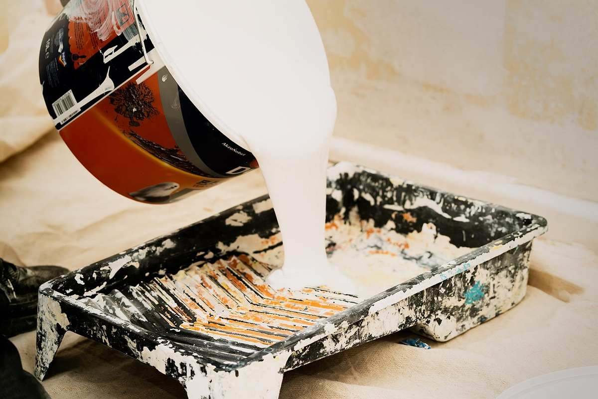 Versement de peinture blanche depuis un pot dans un bac de peintre maculé de résidus colorés