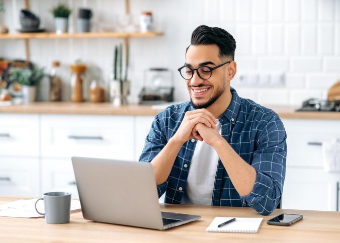Un jeune homme souriant, lunettes, chemise à carreaux, devant son ordinateur